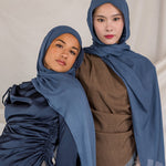 Two women wearing blue hijabs and gray pants against a plain background