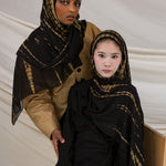 Two models wearing the Turmeric Ripple hijab in colors ranging from black to golden yellow, in a tie-dye pattern.