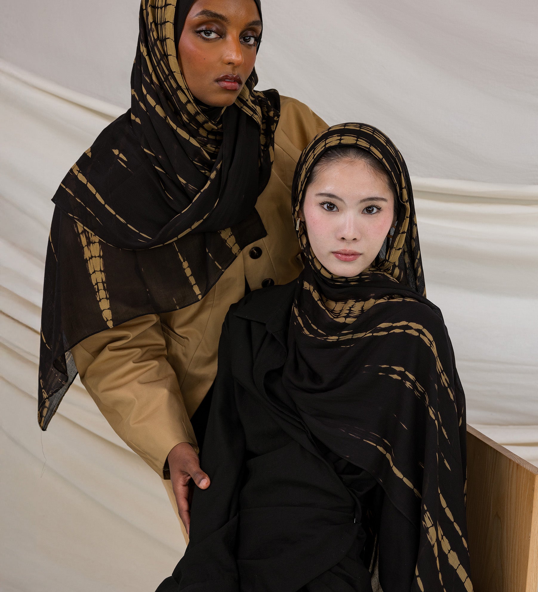 Two models wearing the Turmeric Ripple hijab in colors ranging from black to golden yellow, in a tie-dye pattern.