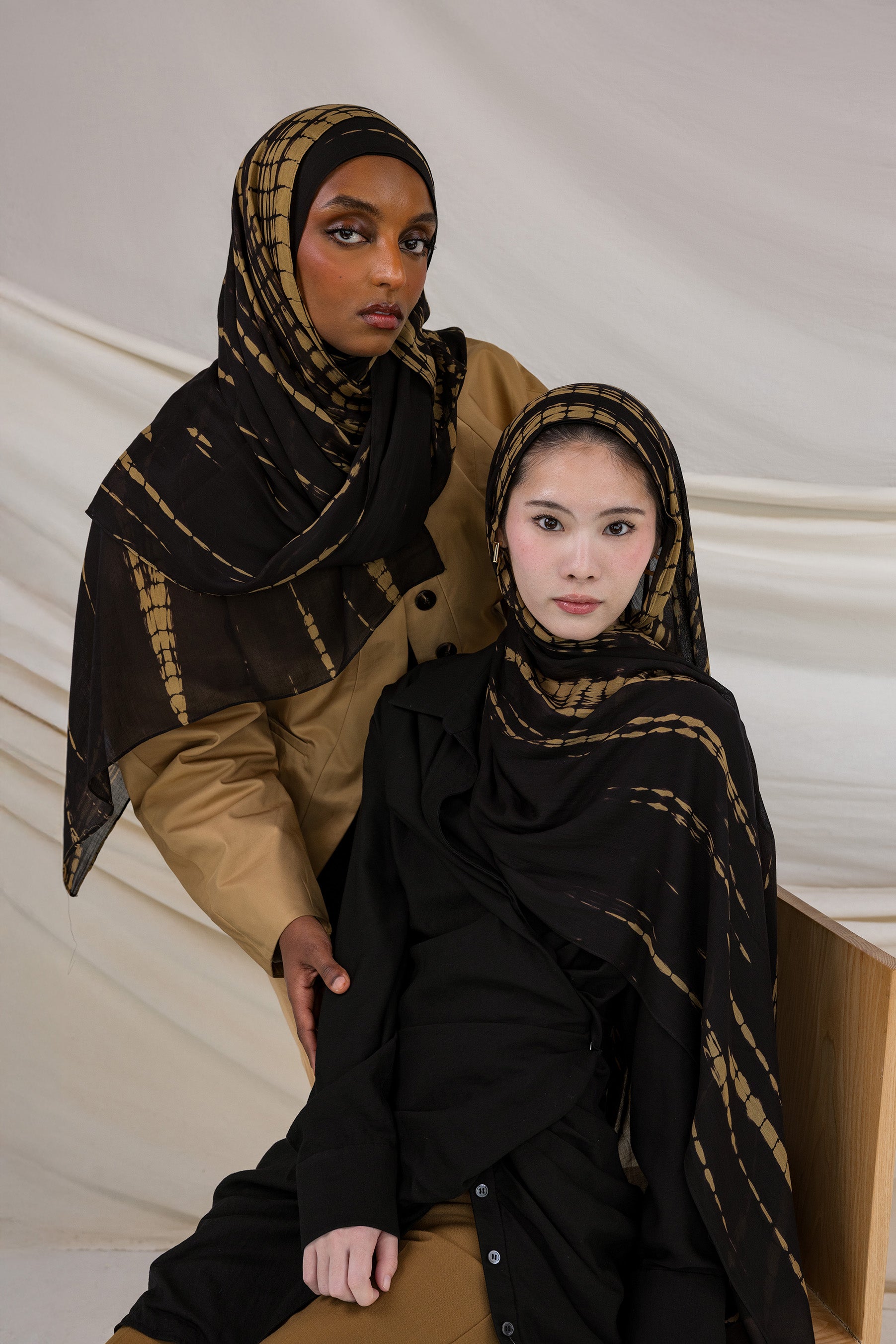 Two models wearing the Turmeric Ripple hijab in colors ranging from black to golden yellow, in a tie-dye pattern.