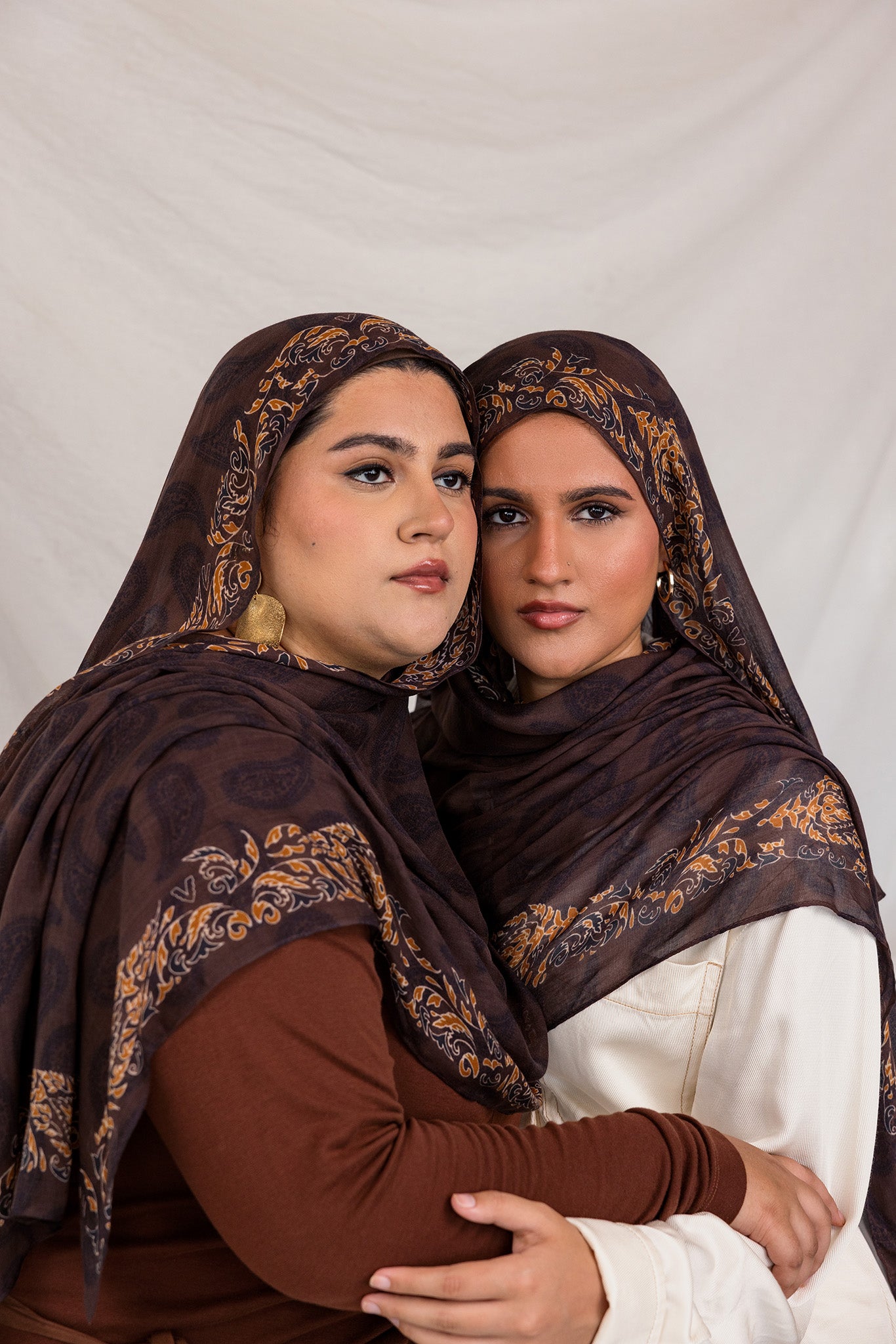 Sister models in Chocolate Paisley hijabs holding each other posed together in studio light.