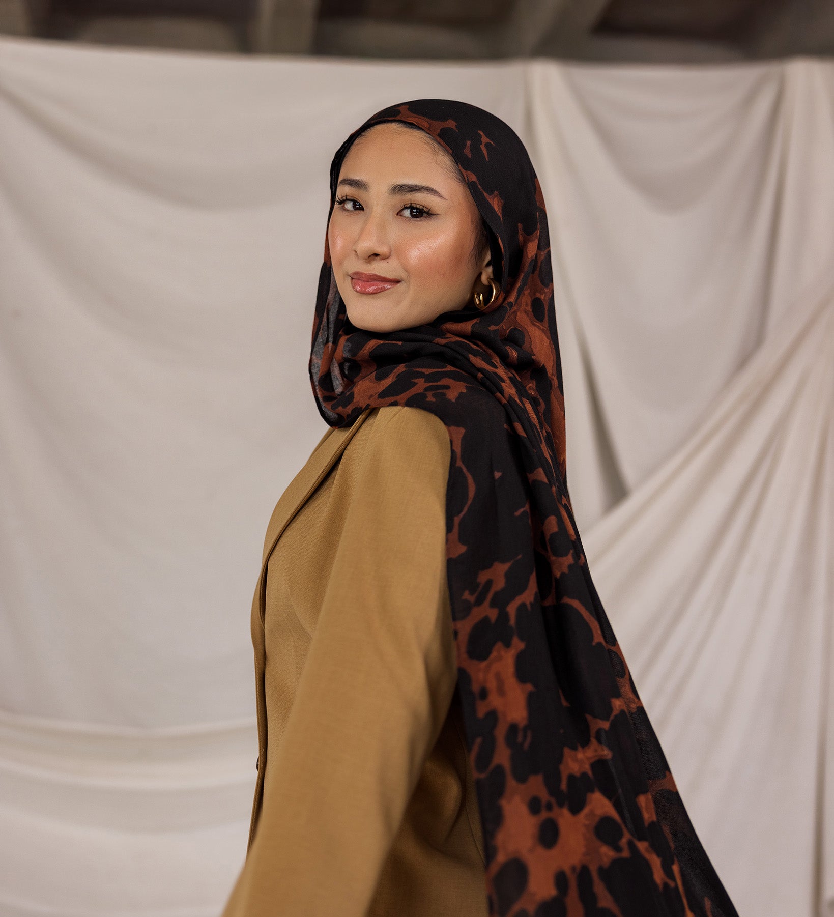 Woman wearing a tortoiseshell patterned hijab and beige top against a plain background