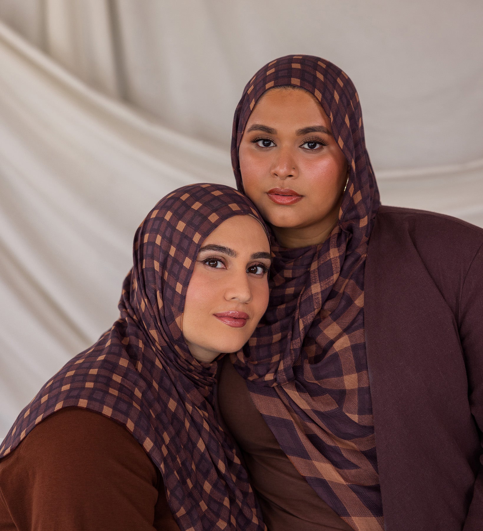Two women wearing matching brown and purple checkered plaid print hijabs against a plain background