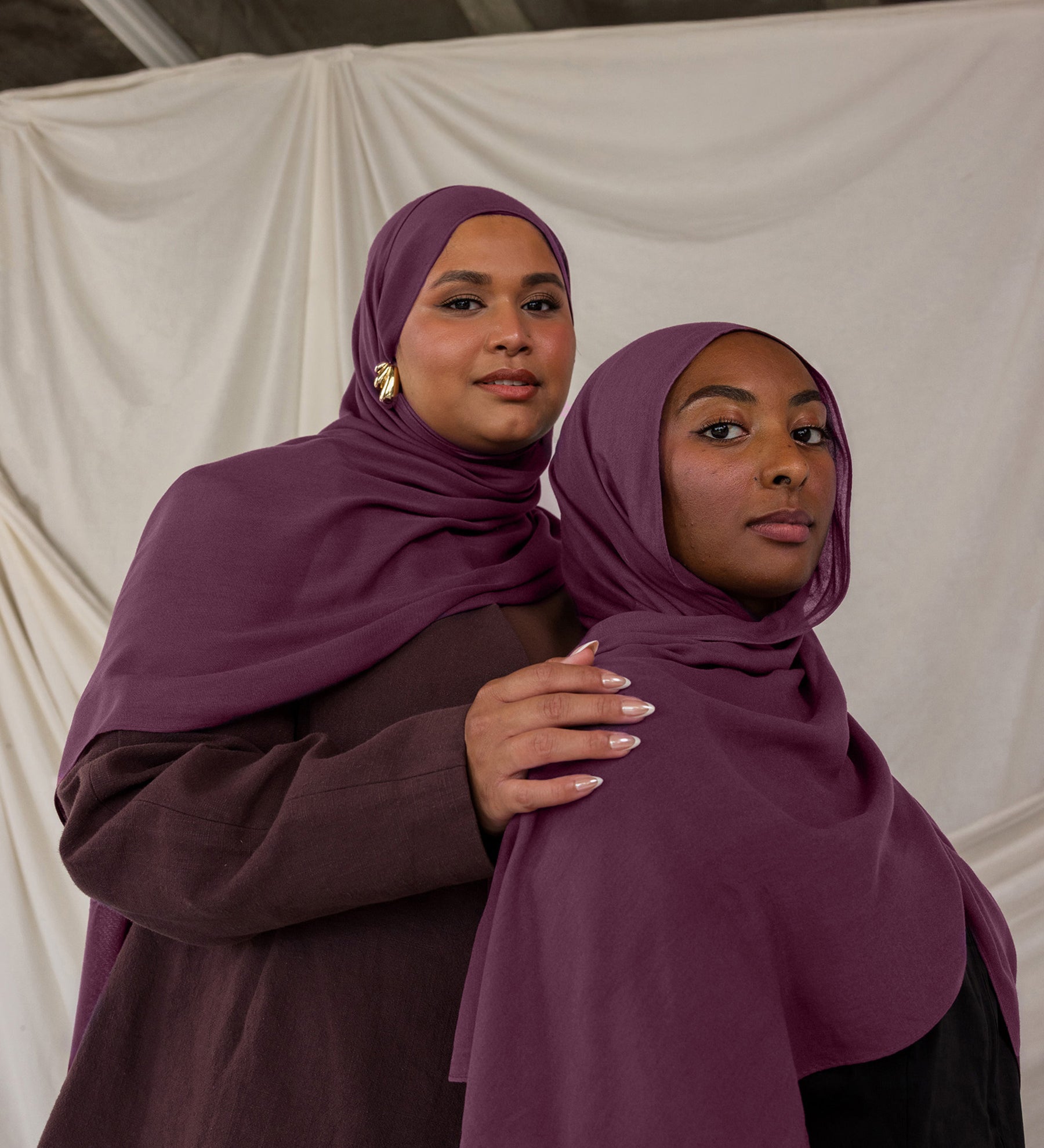 Two women wearing purple red toned hijabs standing close to each other against a plain background.