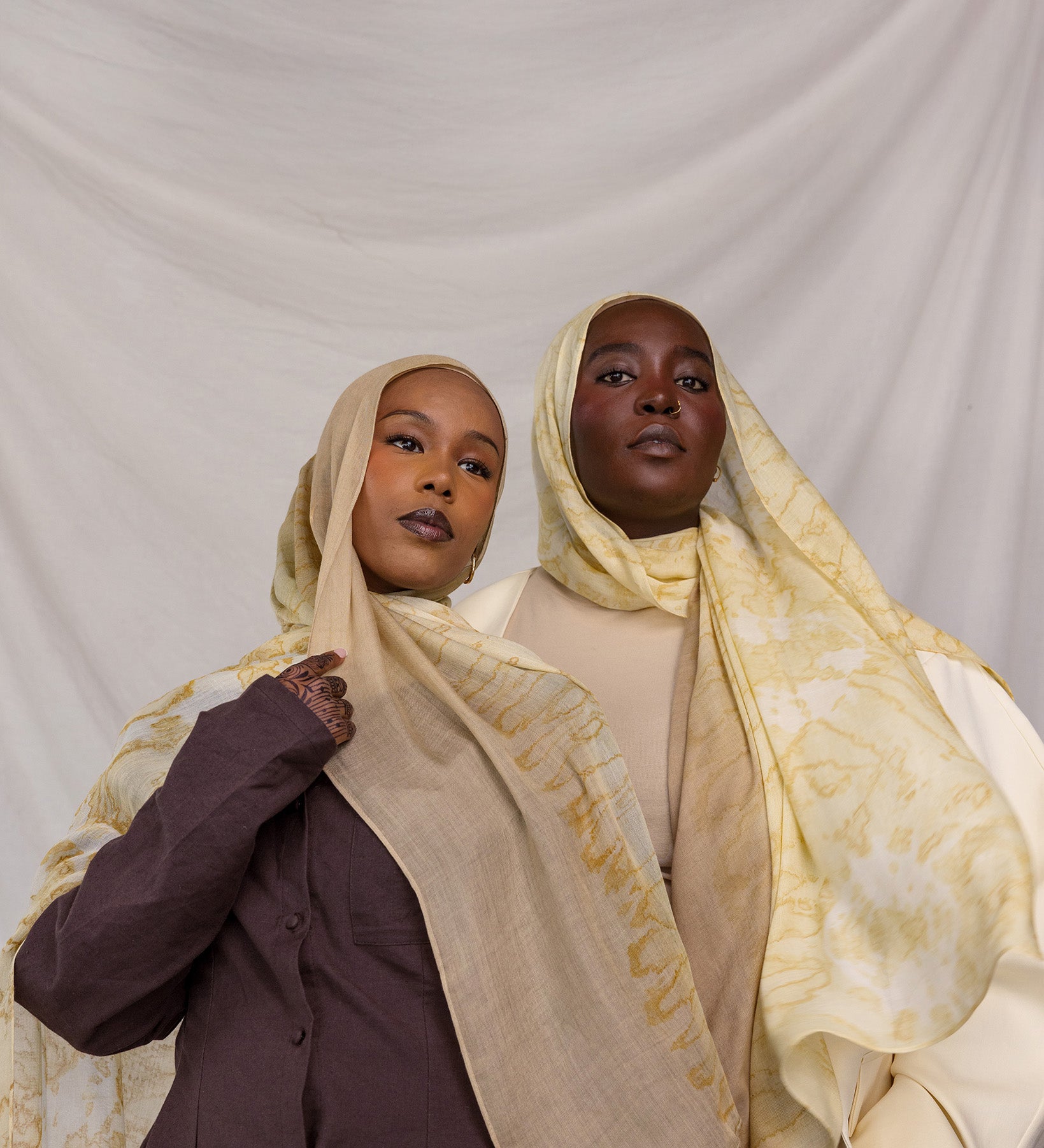 Two women wearing yellow and tan printed headscarves against a plain background