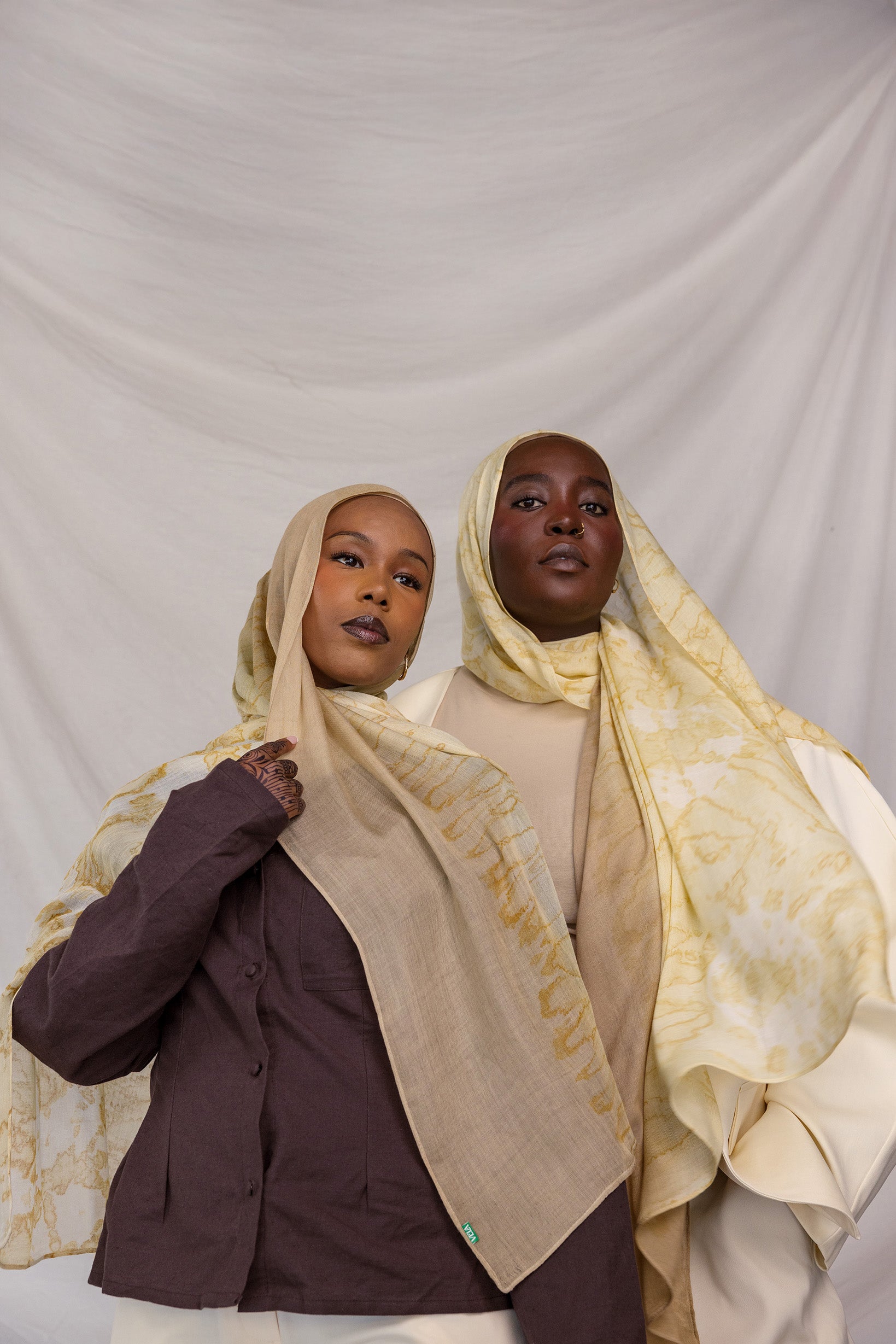 Two women wearing yellow and tan printed headscarves against a plain background