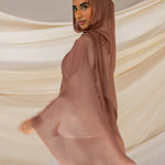 A person wearing a brown hijab with reddish undertones, standing with their back towards the camera with a draped fabric in the background.