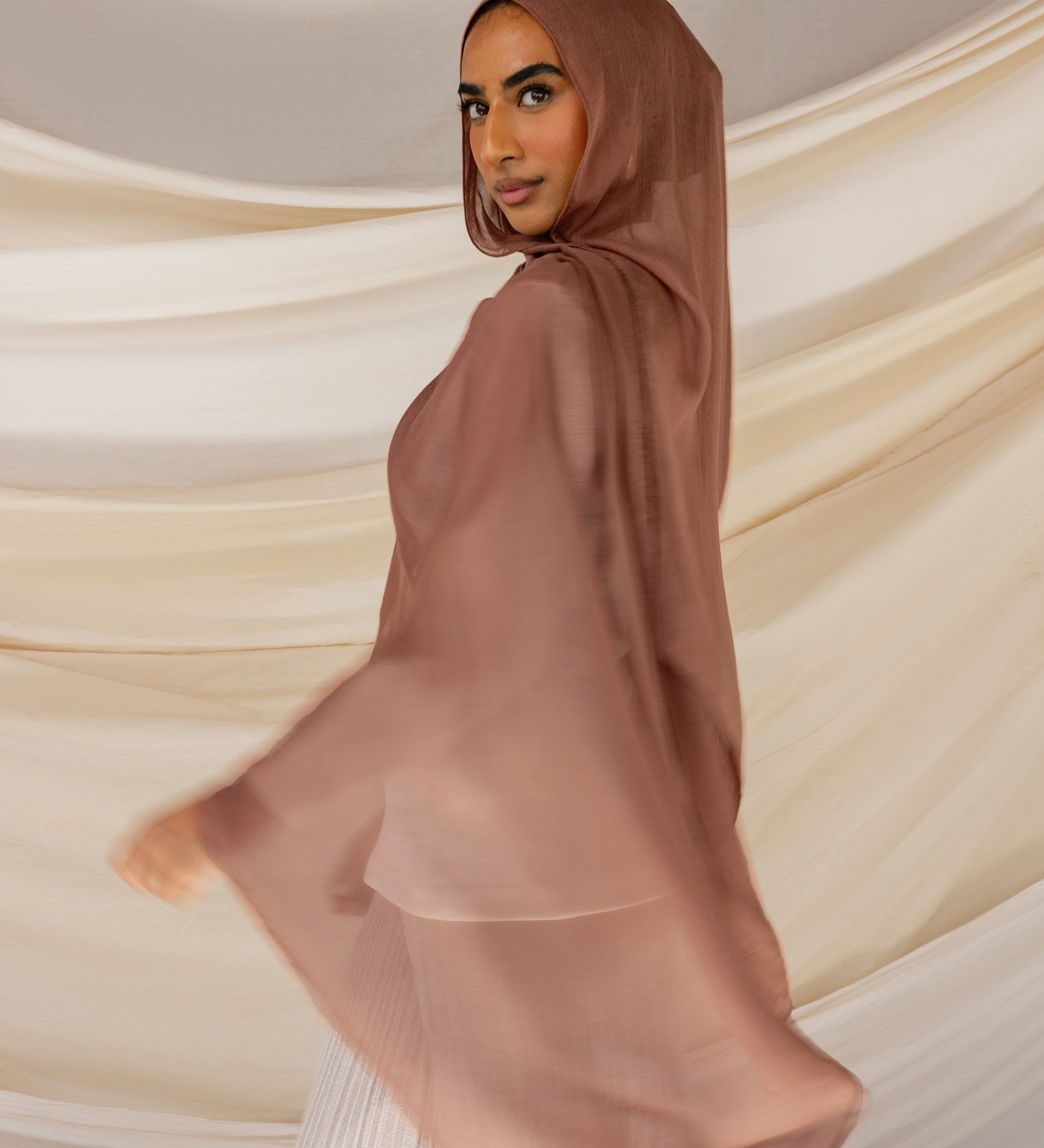 A person wearing a brown hijab with reddish undertones, standing with their back towards the camera with a draped fabric in the background.