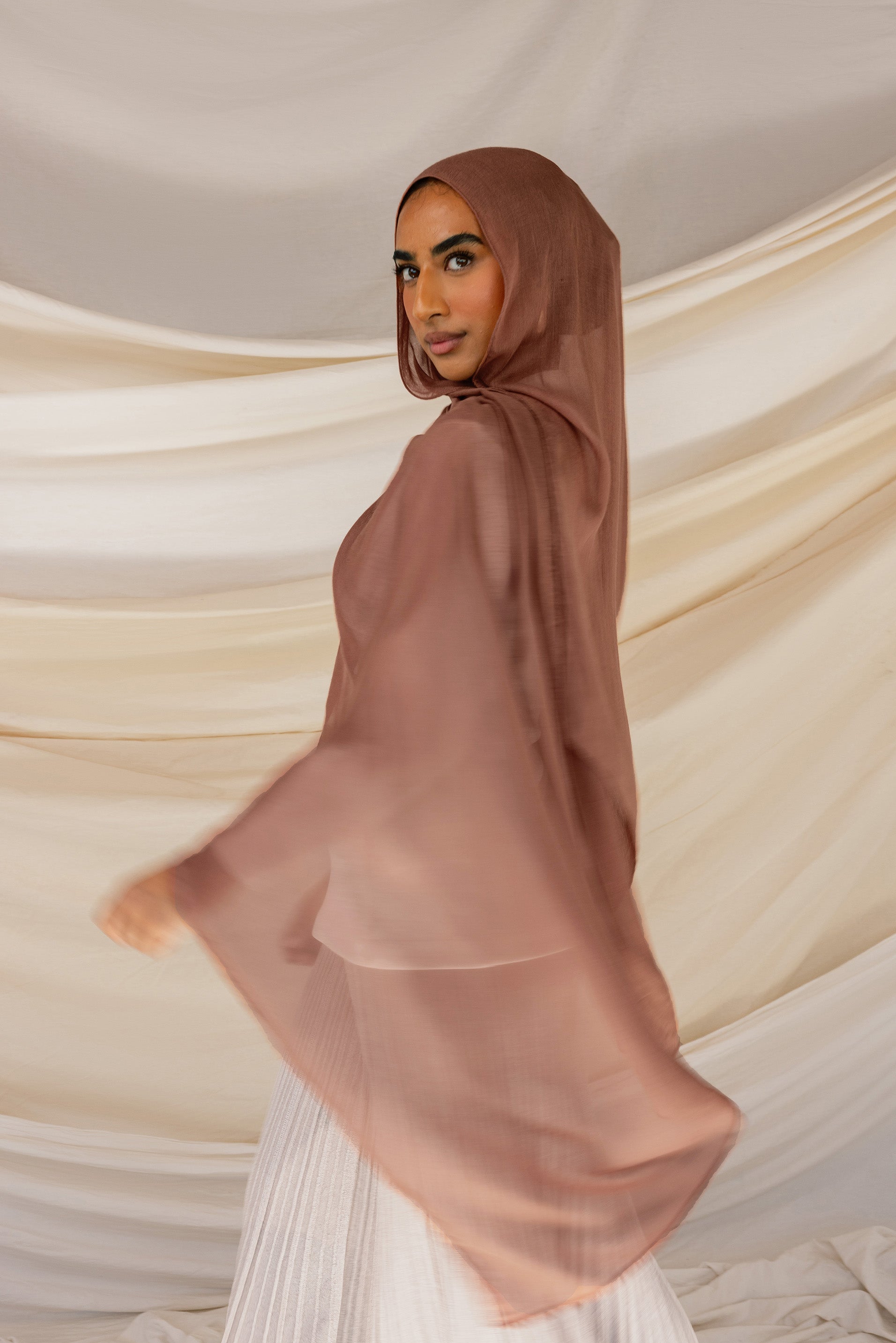 A person wearing a brown hijab with reddish undertones, standing with their back towards the camera with a draped fabric in the background.