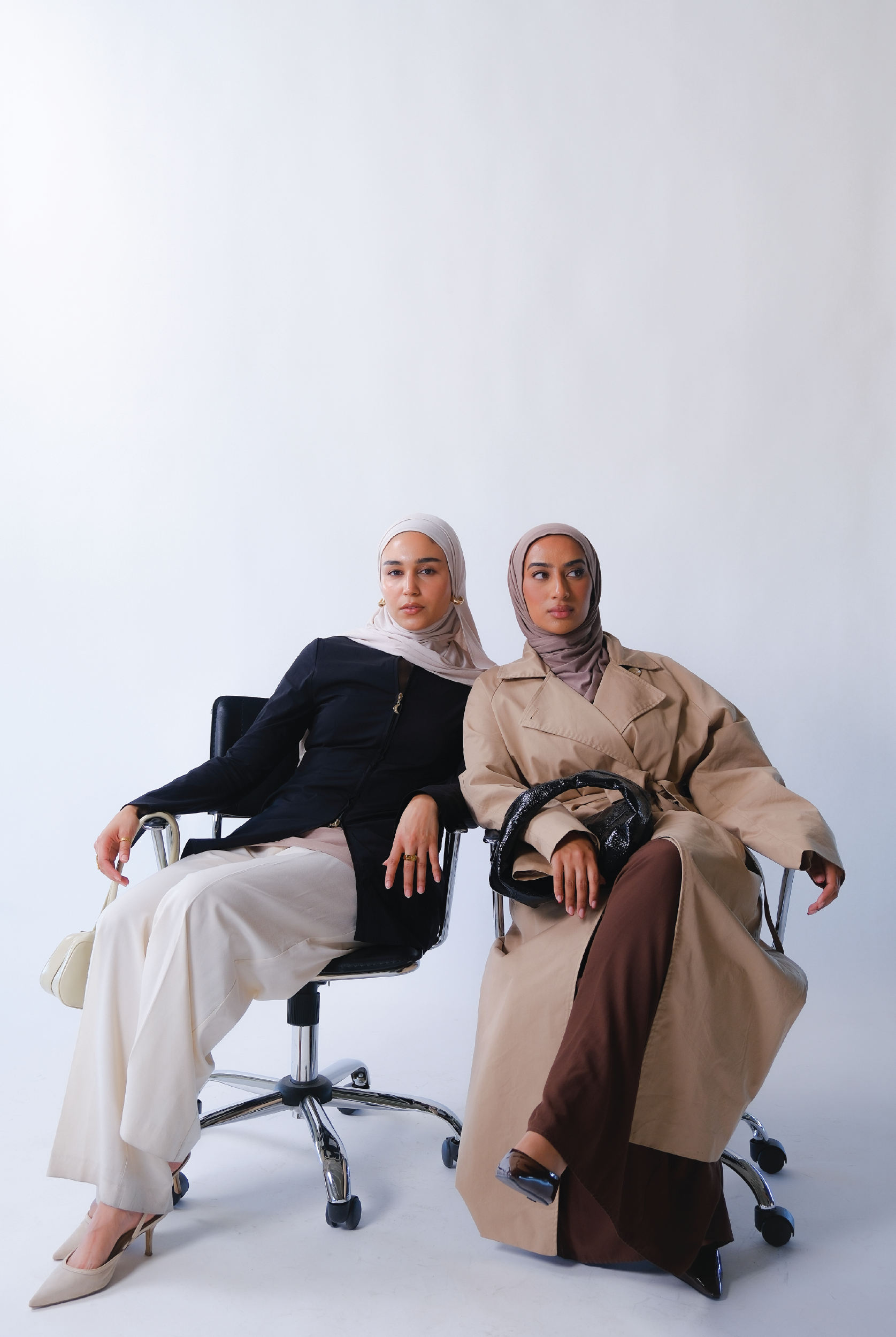 Two women wearing hijabs sitting side by side against a plain background