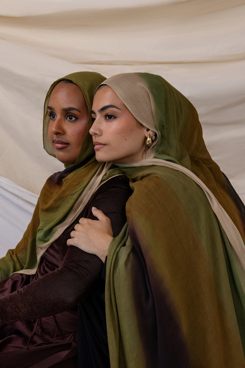 Two women wearing green headscarves and brown dresses against a beige background