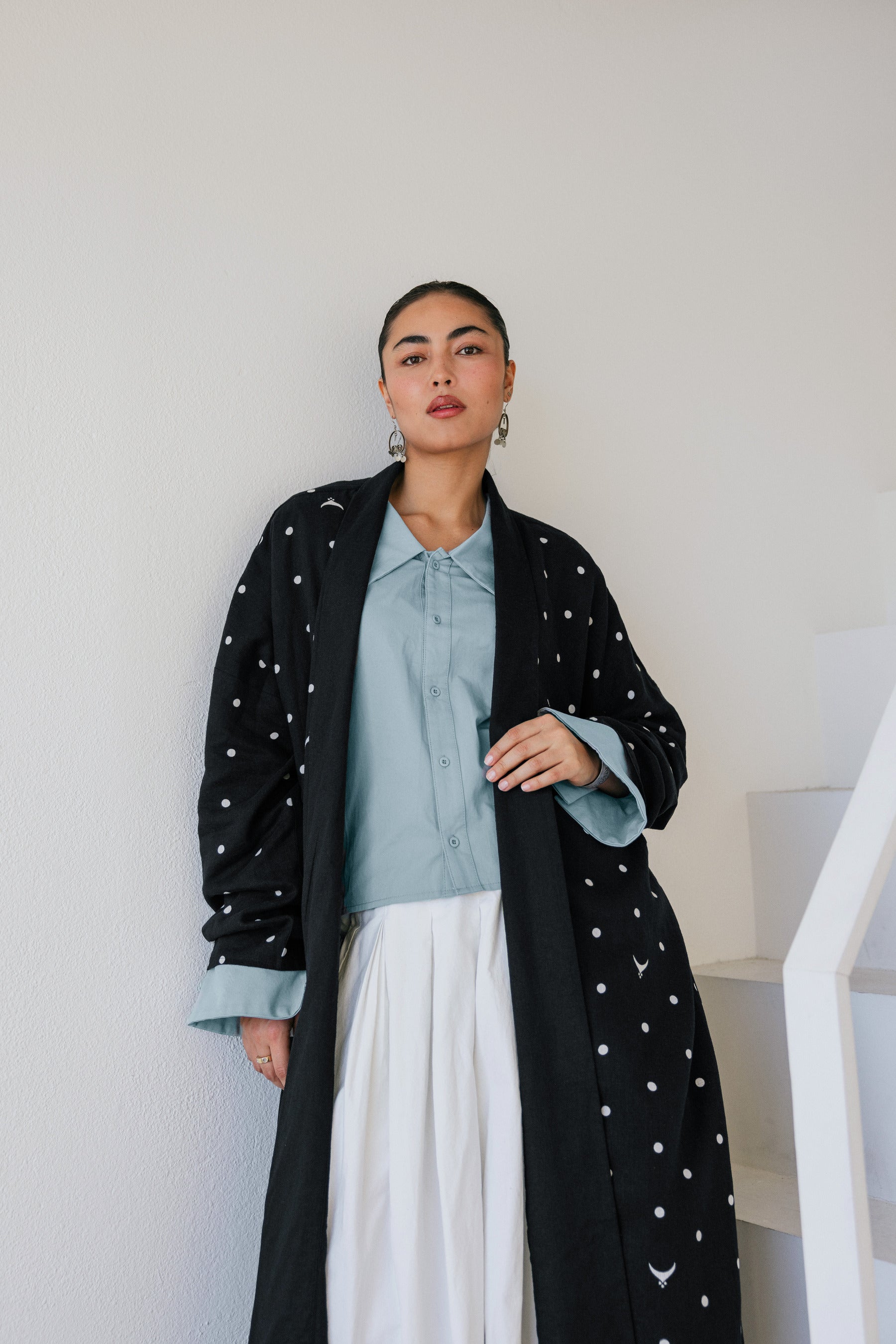Woman wearing a black abaya with white polka dots, light blue shirt, and white skirt against a white wall.