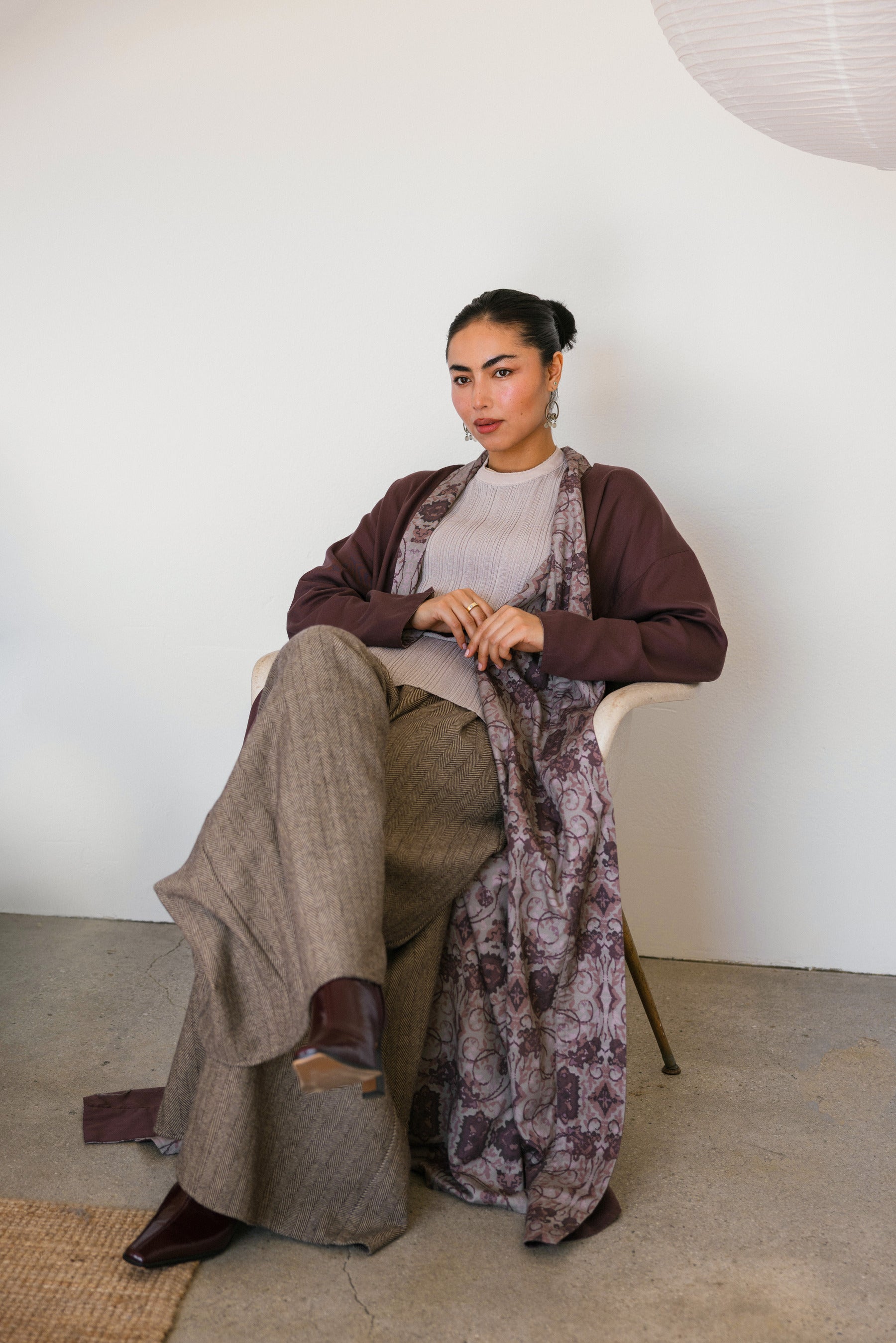 Woman sitting on a chair wearing a patterned abaya and brown pants against a white wall.