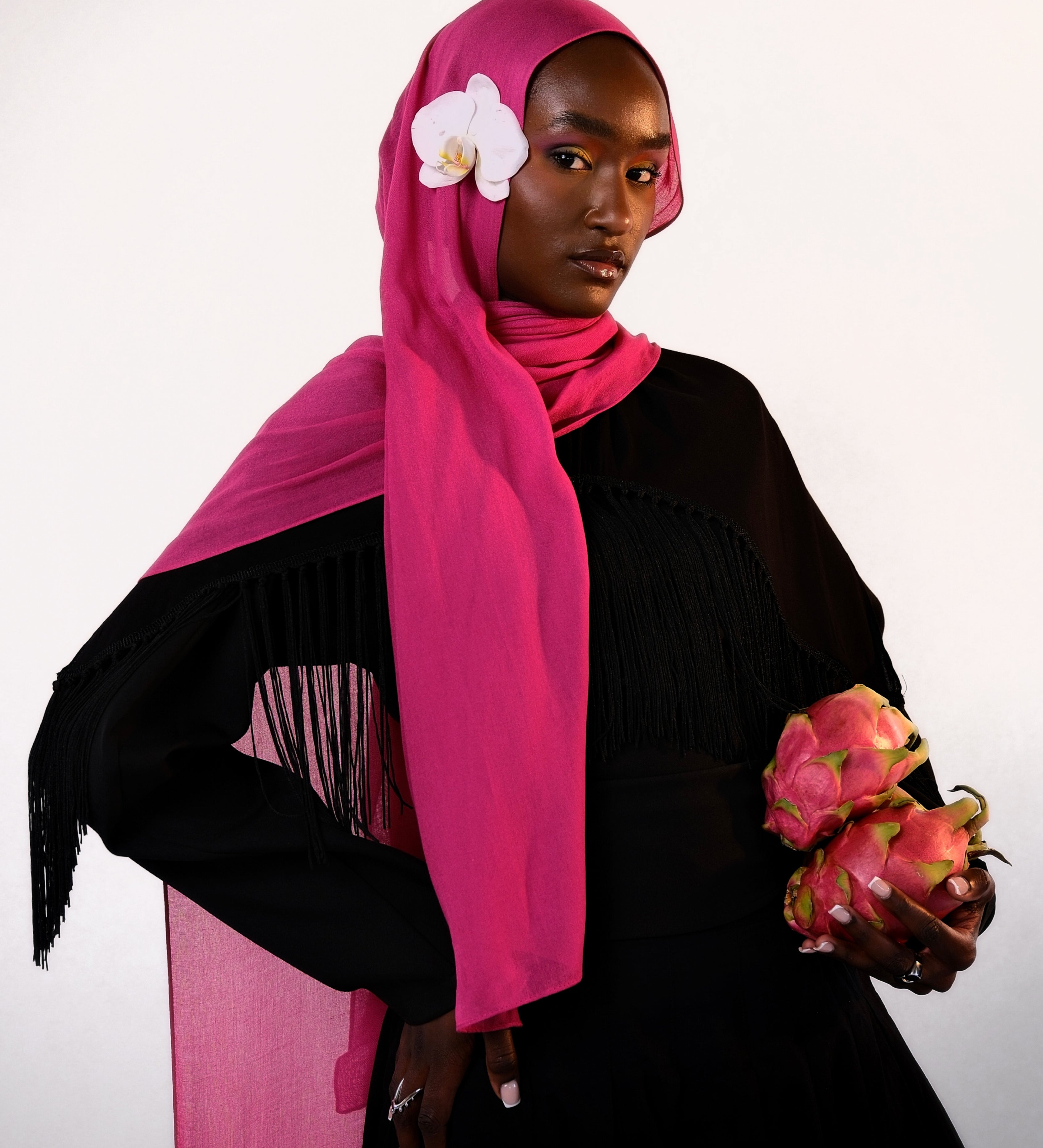 Woman wearing a bright pink hijab with a white flower, holding dragon fruit, on a light gray background