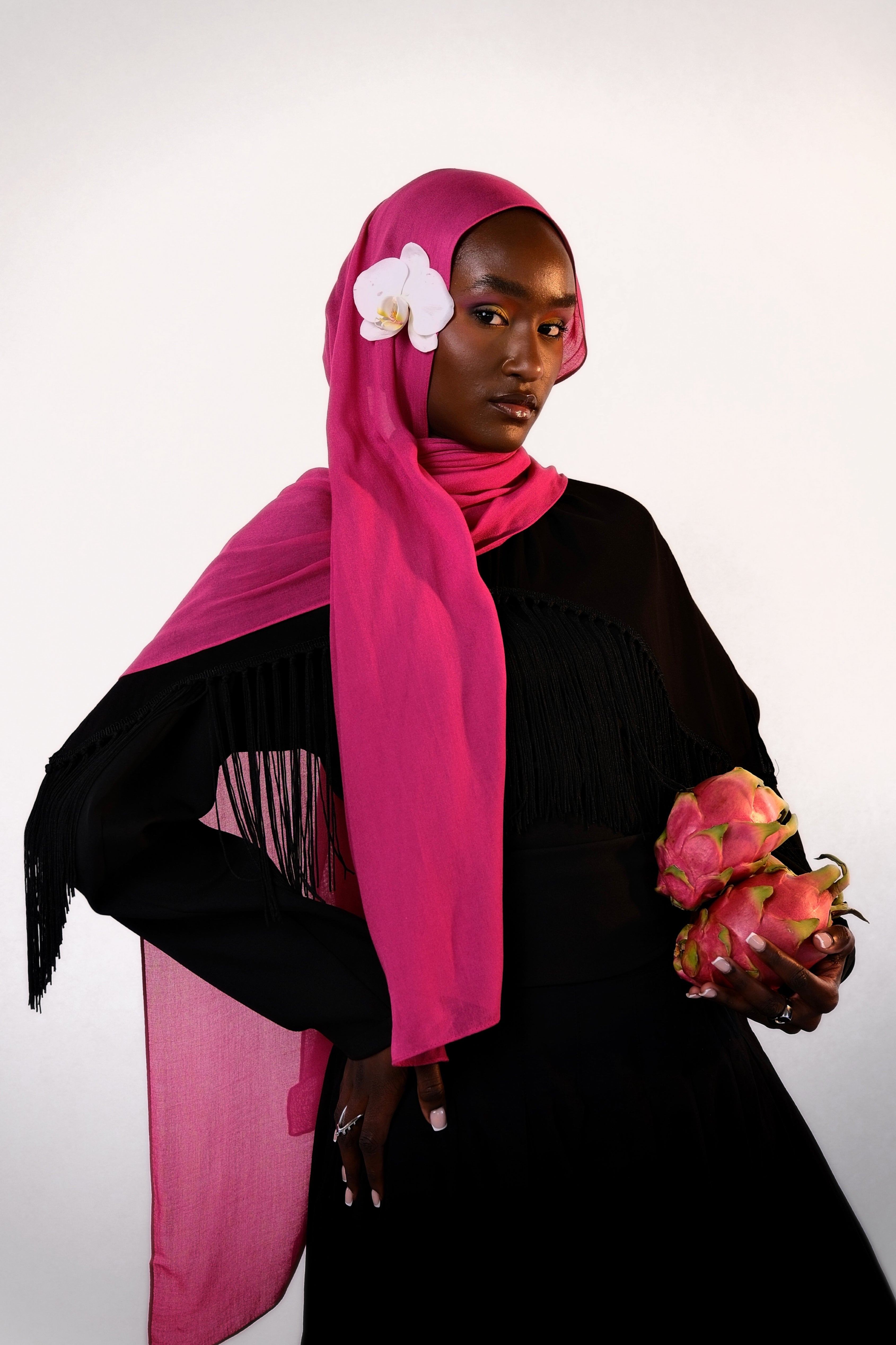 Woman wearing a bright pink hijab with a white flower, holding dragon fruit, on a light gray background