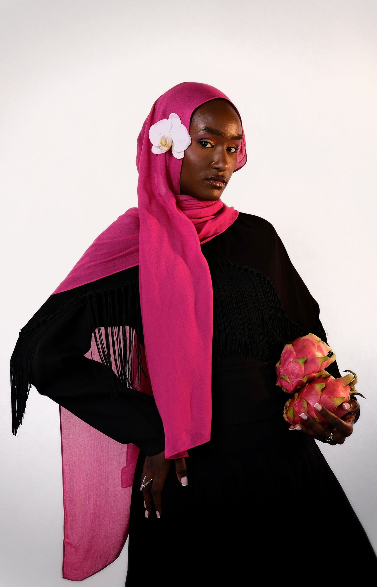 Woman wearing a bright pink hijab with a white flower, holding dragon fruit, on a light gray background