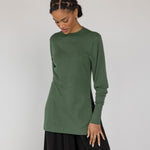 Woman wearing a green long-sleeve top and black pants against a white background