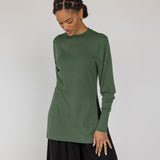 Woman wearing a green long-sleeve top and black pants against a white background