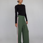 Woman wearing a black top and green wide-leg pants with pink side panels on a gray background