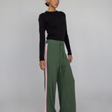 Woman wearing a black top and green wide-leg pants with pink side panels on a gray background