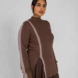 Woman wearing a brown outfit with a high collar and side slits on a plain background