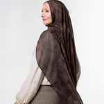 A woman wearing a brown patterned hijab and a cream blouse with a brown skirt, shown from a side profile against a light gray studio background.