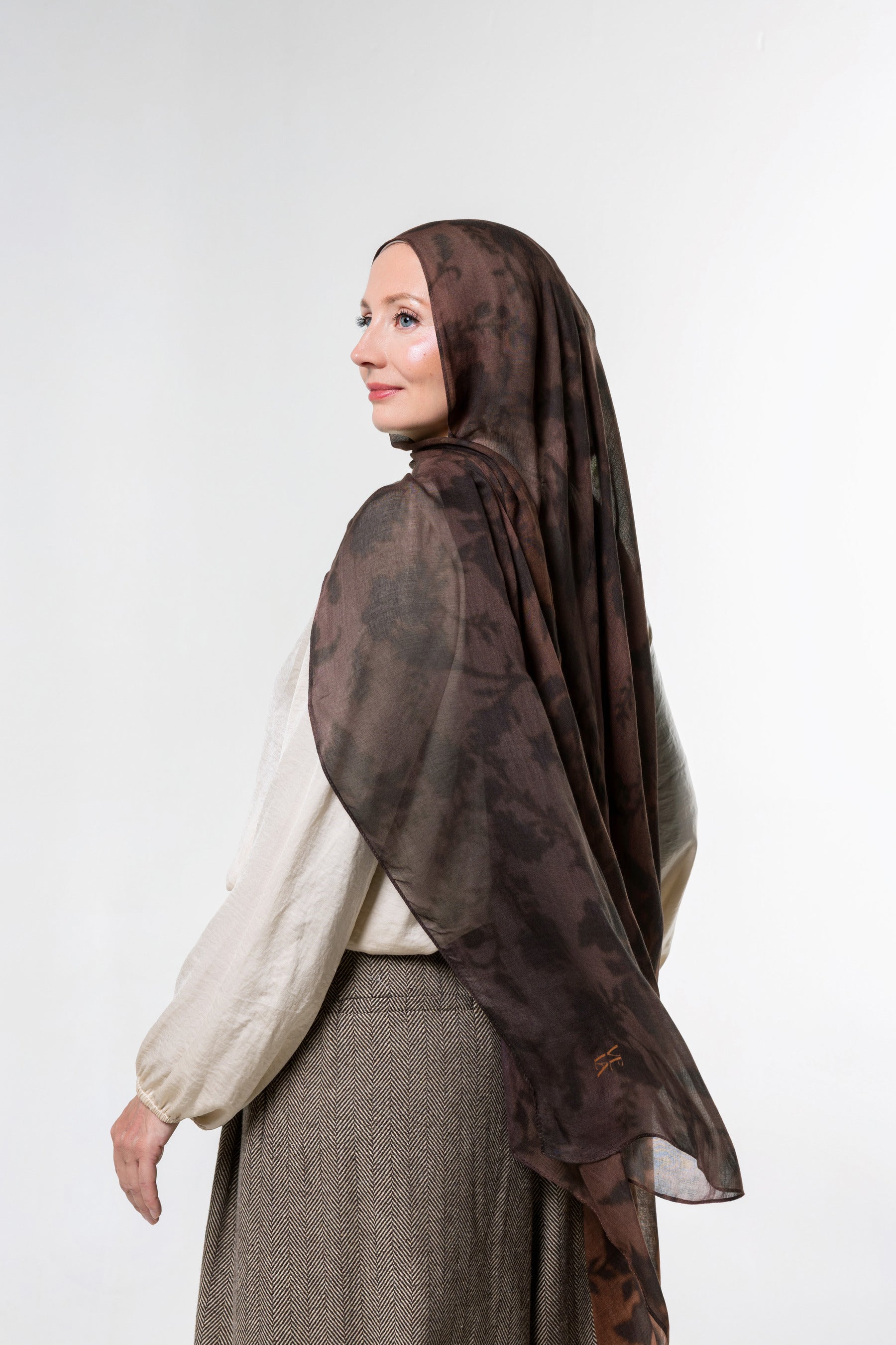 A woman wearing a brown patterned hijab and a cream blouse with a brown skirt, shown from a side profile against a light gray studio background.