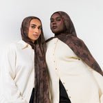 Two women modeling matching brown and black patterned hijabs and cream button-up shirts against a plain light gray background.