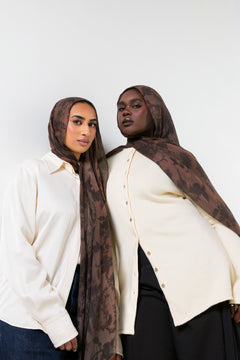 Two women modeling matching brown and black patterned hijabs and cream button-up shirts against a plain light gray background.