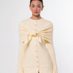 A model wearing a pale yellow knit cardigan with a large front bow detail and button-up front, paired with brown pants, against a plain light gray background.