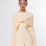 A model wearing a pale yellow knit cardigan with a large front bow detail and button-up front, paired with brown pants, against a plain light gray background.