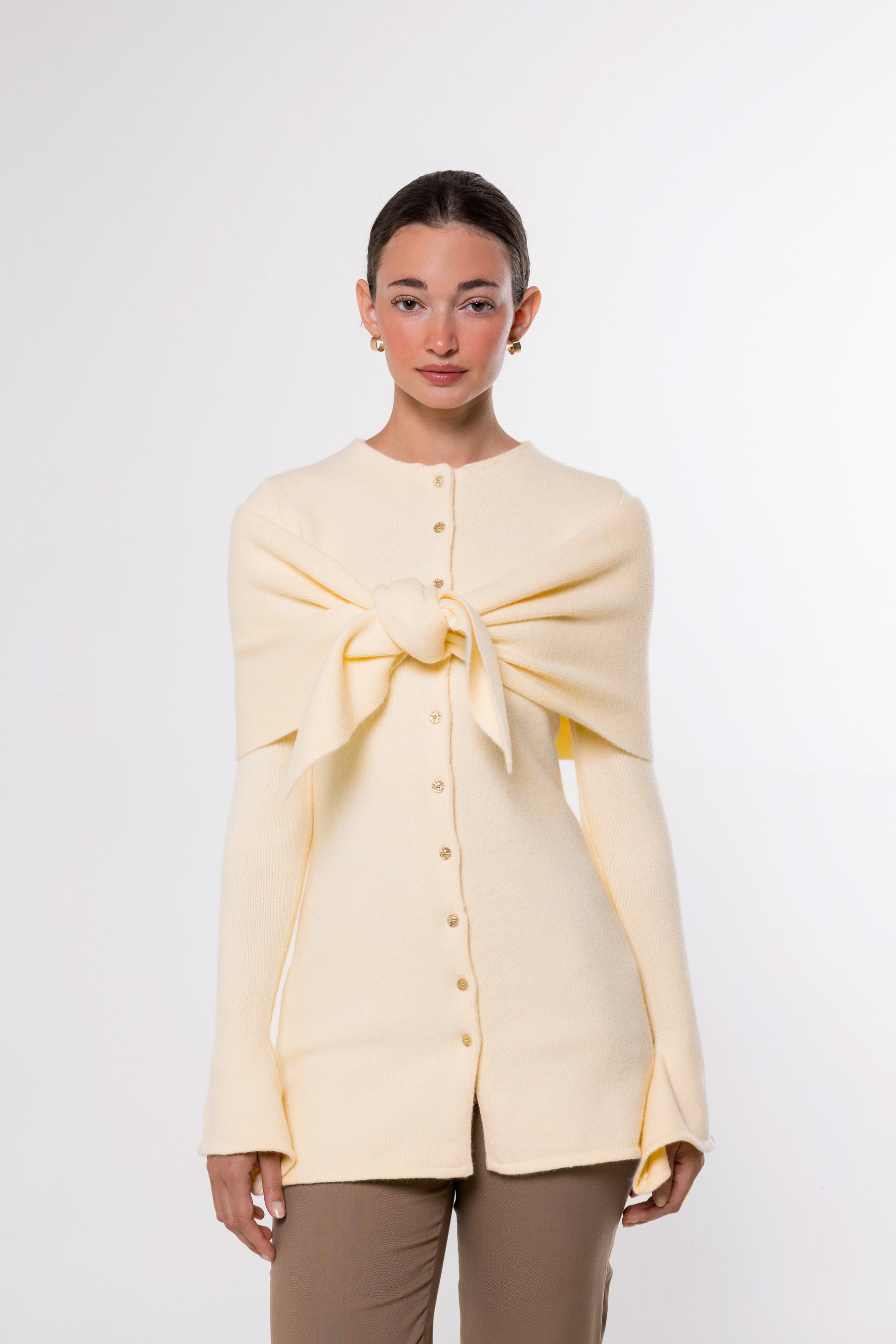 A model wearing a pale yellow knit cardigan with a large front bow detail and button-up front, paired with brown pants, against a plain light gray background.