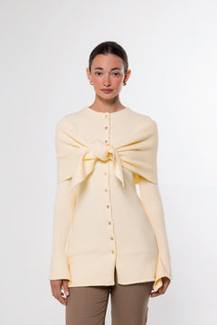 A model wearing a pale yellow knit cardigan with a large front bow detail and button-up front, paired with brown pants, against a plain light gray background.