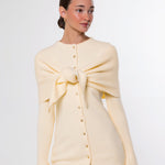 A woman modeling a cream-colored knit cardigan with a large bow detail at the chest and a button-down front, paired with brown pants, against a plain white studio background.
