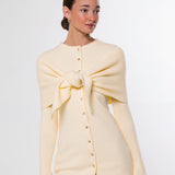 A woman modeling a cream-colored knit cardigan with a large bow detail at the chest and a button-down front, paired with brown pants, against a plain white studio background.
