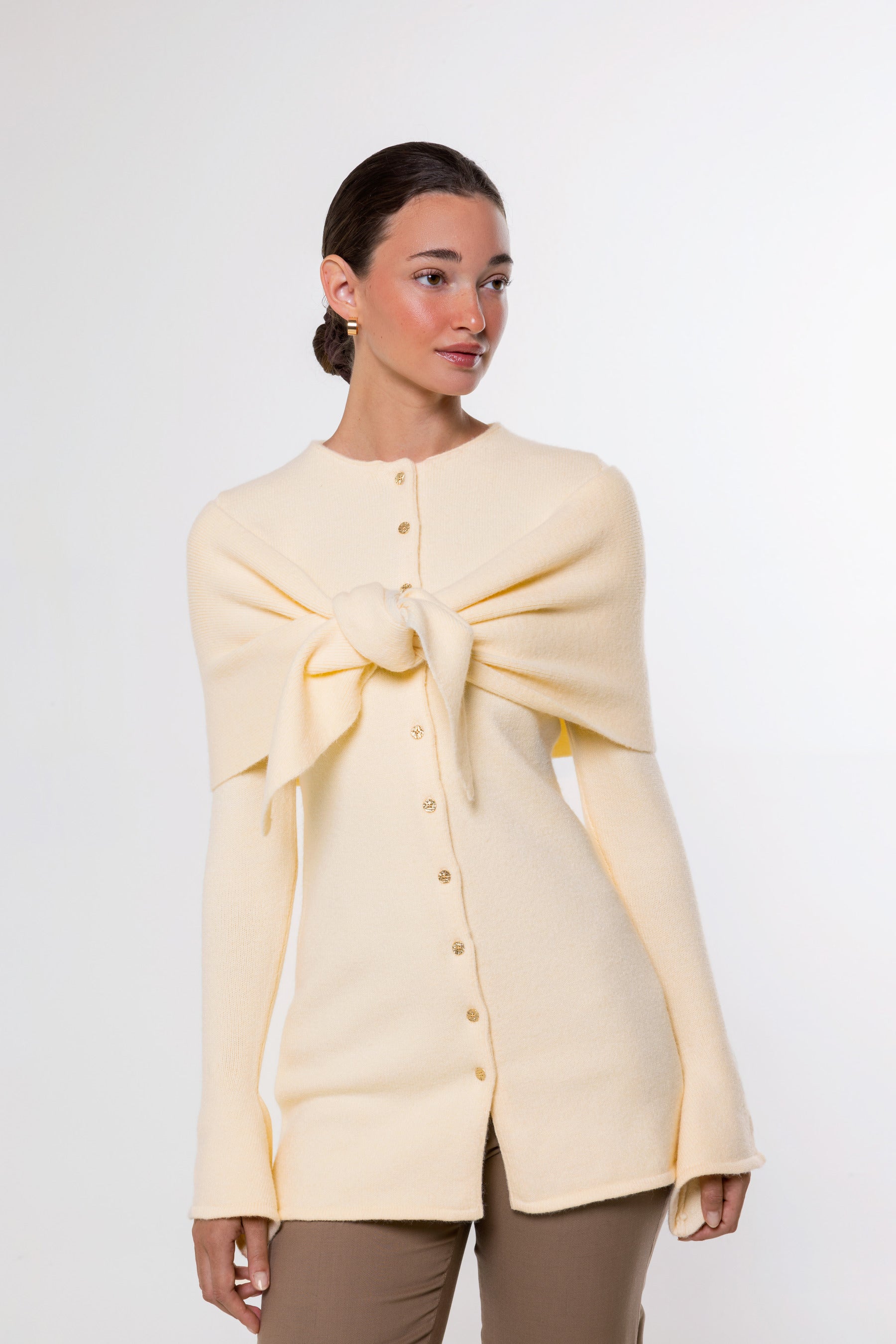 A woman modeling a cream-colored knit cardigan with a large bow detail at the chest and a button-down front, paired with brown pants, against a plain white studio background.