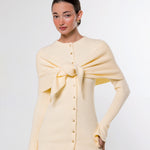 A woman modeling a pale cream knit cardigan with a large front bow detail and button closure, paired with taupe pants, against a plain light gray studio background.
