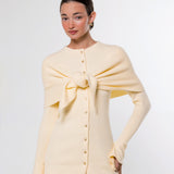 A woman modeling a pale cream knit cardigan with a large front bow detail and button closure, paired with taupe pants, against a plain light gray studio background.