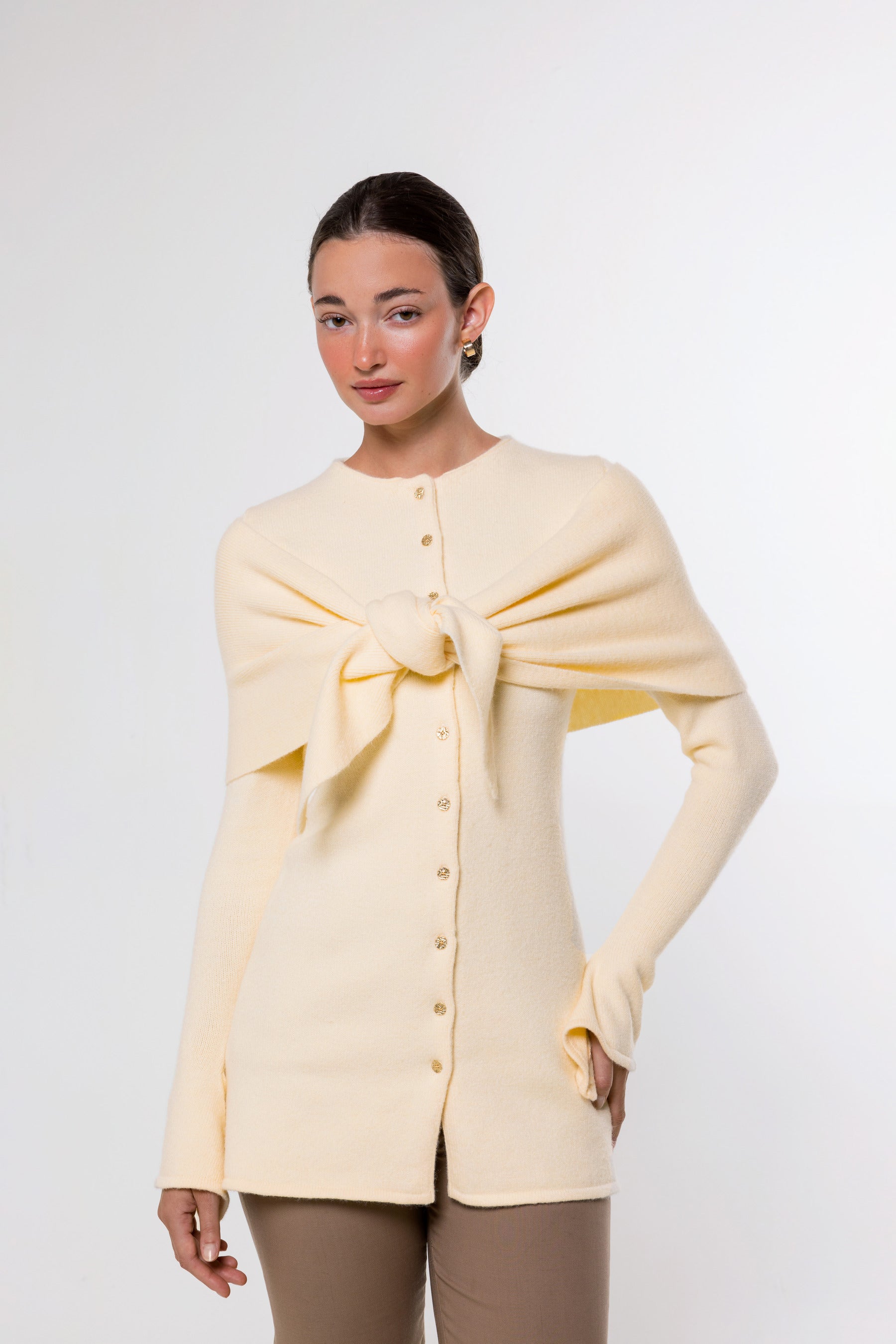 A woman modeling a pale cream knit cardigan with a large front bow detail and button closure, paired with taupe pants, against a plain light gray studio background.