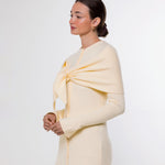 A woman modeling a pale cream knit wrap or shawl draped over her shoulders and tied at the chest, paired with taupe leggings, against a plain light gray studio background.