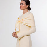 A woman modeling a pale cream knit wrap or shawl draped over her shoulders and tied at the chest, paired with taupe leggings, against a plain light gray studio background.