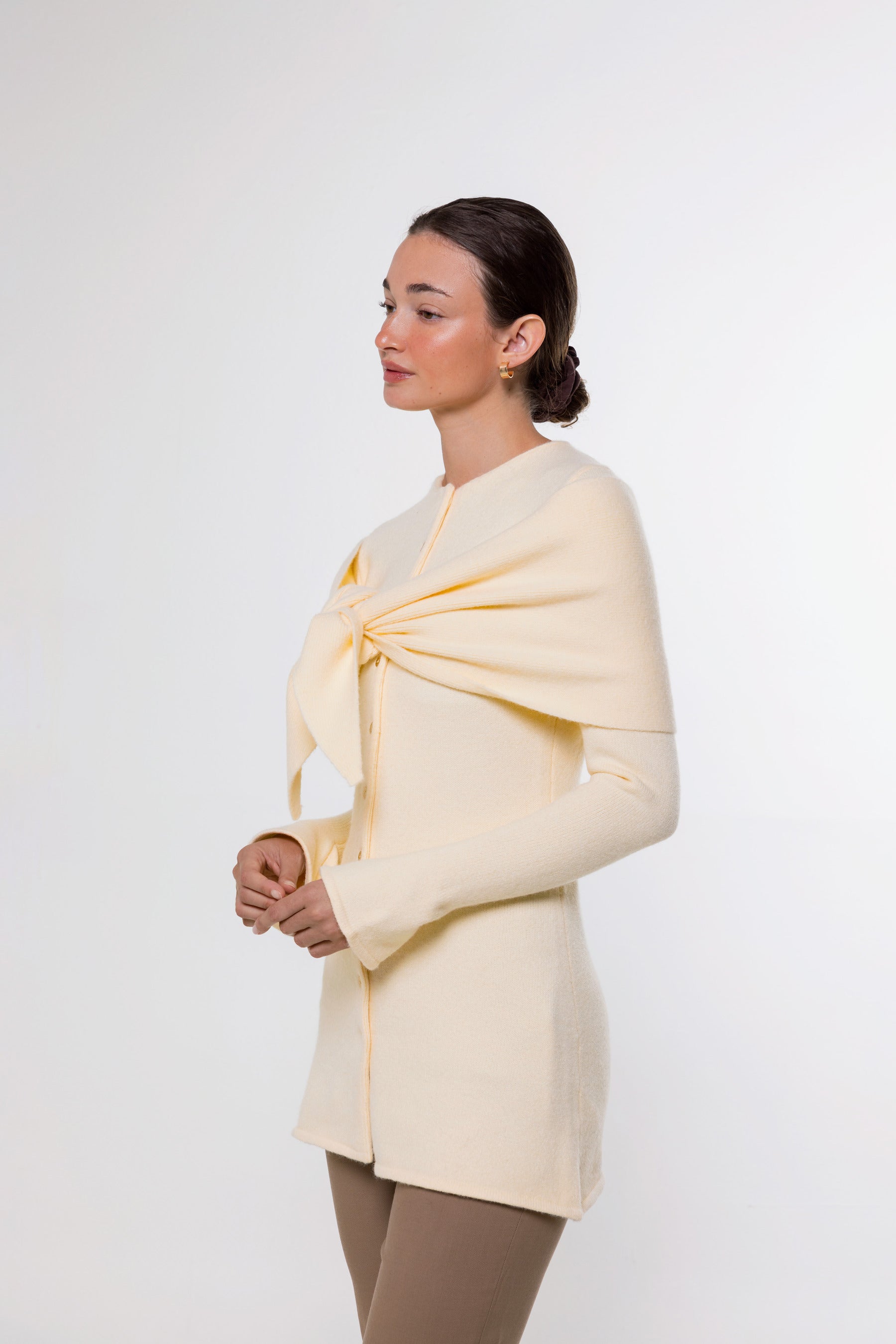 A woman modeling a pale cream knit wrap or shawl draped over her shoulders and tied at the chest, paired with taupe leggings, against a plain light gray studio background.