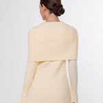 A woman shown from the back wearing a light cream or pale yellow knit top with a wide draped yoke and long sleeves, paired with taupe pants, against a plain light gray studio background.