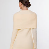 A woman shown from the back wearing a light cream or pale yellow knit top with a wide draped yoke and long sleeves, paired with taupe pants, against a plain light gray studio background.