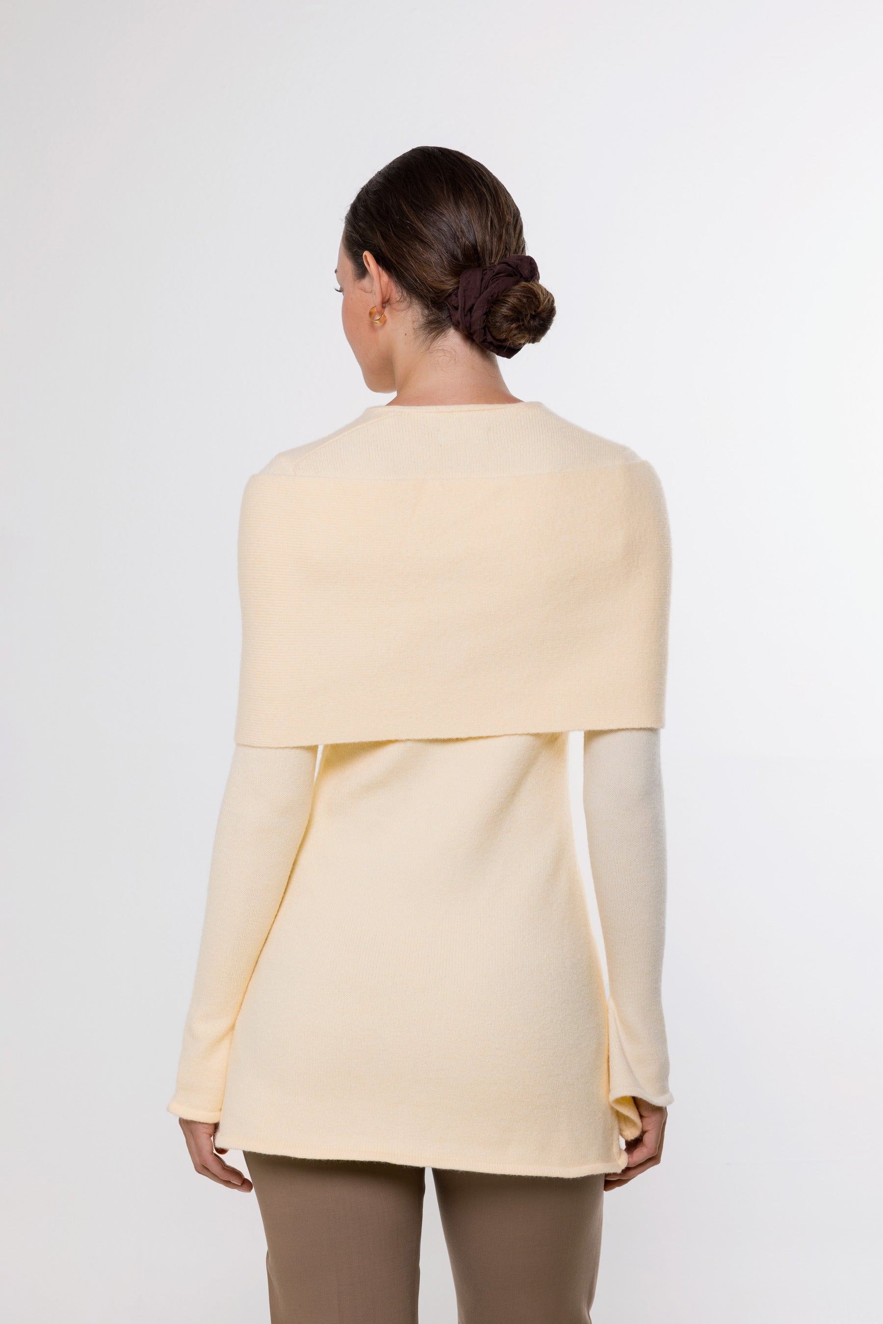 A woman shown from the back wearing a light cream or pale yellow knit top with a wide draped yoke and long sleeves, paired with taupe pants, against a plain light gray studio background.