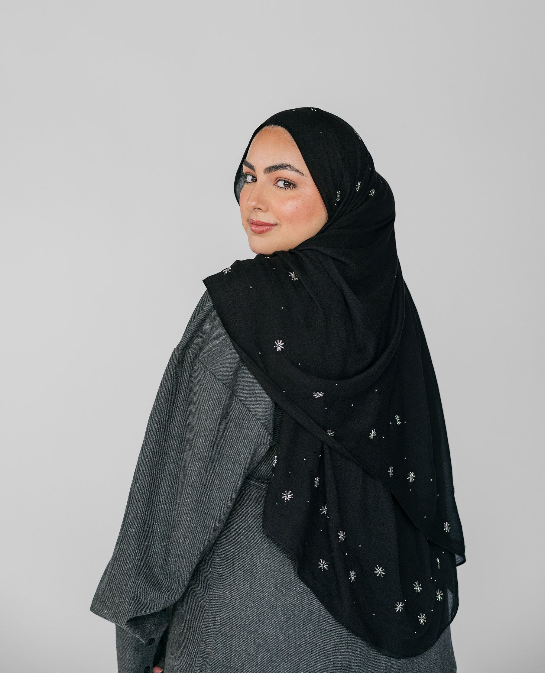Woman wearing a black hijab with white beadeed patterns on a gray background