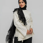 A woman wearing a black hijab with a dramatic cascading ruffle, a cream-colored long-sleeve blouse, and black wide-leg trousers, photographed against a solid light gray studio background.