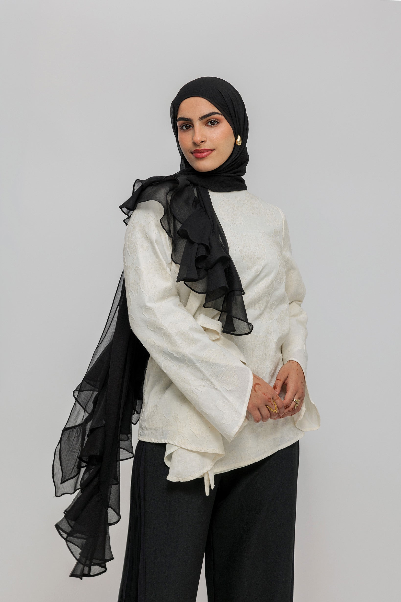 A woman wearing a black hijab with a dramatic cascading ruffle, a cream-colored long-sleeve blouse, and black wide-leg trousers, photographed against a solid light gray studio background.