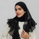 A woman wearing a black sheer ruffled hijab and a cream-colored top, posing against a solid light gray background.