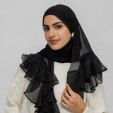 A woman wearing a black sheer ruffled hijab and a cream-colored top, posing against a solid light gray background.